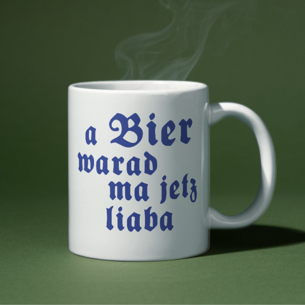 Weiße Keramiktasse „A Bier warad ma a liaba“ auf einem bayerischen Frühstückstisch mit Brezen und Maßkrug