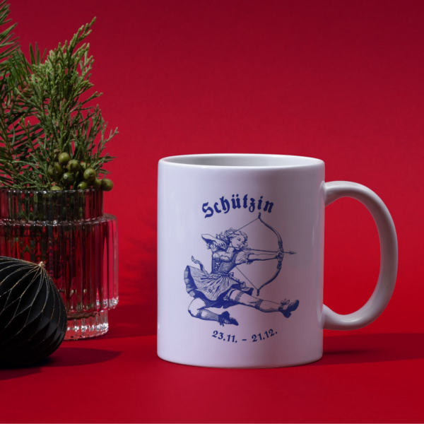 Bayerische Keramiktasse mit Schütze-Motiv auf einem Tisch, umgeben von winterlicher Weihnachtsdeko und einer Tasse Tee.